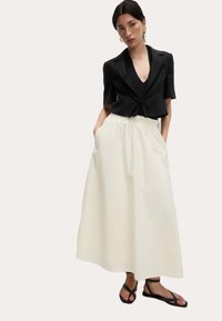 Schwarze, taillierte Blazerjacke mit kurzen Ärmeln und einer Schleifenfront, kombiniert mit einem weißen, fließenden, knöchellangen Rock. Sandalen mit dünnen Riemen.