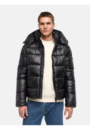 Jeune homme portant une veste matelassée noire brillante avec capuche, un pull crème et un jean bleu, debout devant un fond clair uni.