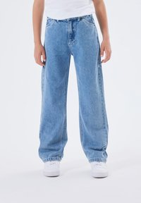 Ljusblå, vida jeans med elastisk avslut vid anklarna, fem fickor och gylf, visat med vita sneakers.