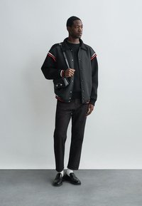 Μαύρο bomber μπουφάν με κόκκινες και λευκές ρίγες, μαύρο πουλόβερ, σκούρα τζιν παντελόνια και μαύρες loafers, αξεσουάρ με μια μικρή τσάντα.