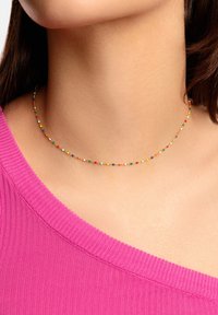 Collar de cuentas coloridas con pequeñas cuentas redondas en varios colores sobre una delgada cadena dorada. Llevado encima de una blusa rosa con estrías.