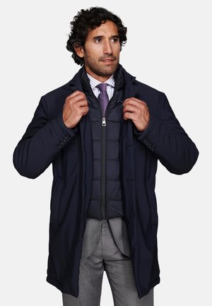Man wearing layered navy coat over black padded jacket, purple tie, white striped shirt, and gray pants, adjusting coat collar.