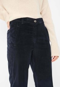 Pantaloni in corduroy blu scuro con una texture liscia, tasche laterali, un bottone in metallo e un design classico a gamba dritta. Il pullover è di colore crema.