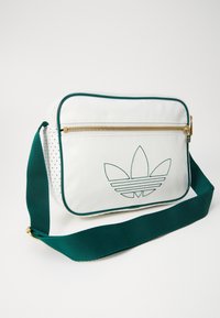 Bílá obdélníková ramenní taška s zeleným lemováním, zlatým zipem, tmavě zeleným nastavitelným popruhem a vyšitým logem Adidas Trefoil na přední kapse.