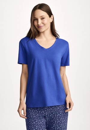 Kvinde smilende, iført en ensfarvet blå V-hals kortærmet skjorte og blå bukser med blomsterprint, stående foran en ensfarvet lys baggrund.