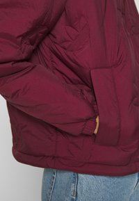 Burgundy puffjacka med subtilt bladmönster, med en quiltad design, elastiska ärmar och sidofickor. Slät, glänsande tyg.