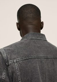 Uomo con capelli corti visto da dietro, che indossa una giacca di jeans nera sbiadita su uno sfondo semplice.