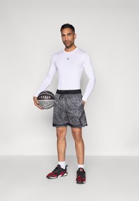 Λευκό πουκάμισο στενής εφαρμογής με μακριά μανίκια, μαύρα και γκρι σορτς με σχέδιο, μαύρα παπούτσια μπάσκετ με κόκκινες λεπτομέρειες, κρατώντας ένα μπάσκετ.