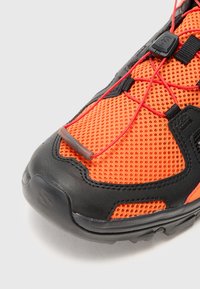 Oransje og svart idrettssko med mesh-overdel, gummisåle og røde elastiske lisser. Har en teksturert overflate og forsterket tåhette.