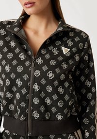 Black zip-up jacket featuring a geometric white pattern and a small triangular silver logo, with textured fabric and ribbed cuffs.