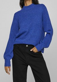Blauwe gebreide trui met een hoge hals en geribde manchetten, gecombineerd met zwarte broek. De trui heeft een ontspannen pasvorm en een zachte textuur.