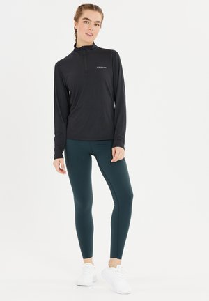 Schwarzes Langarmshirt mit Halb-Reißverschluss und reflektierendem Logo, kombiniert mit dunkelblauen Leggings. Das Modell trägt weiße Sportschuhe.