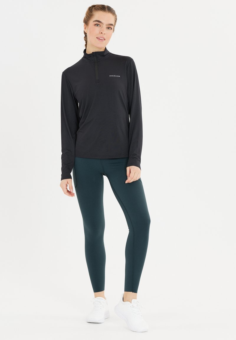 Schwarzes Langarmshirt mit Halb-Reißverschluss und reflektierendem Logo, kombiniert mit dunkelblauen Leggings. Das Modell trägt weiße Sportschuhe.