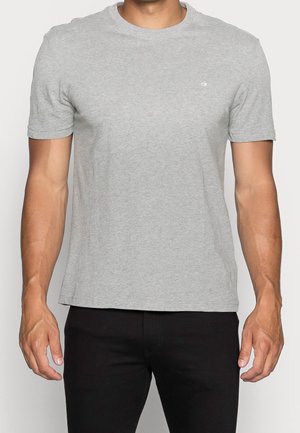 Mand iført lysegrå kortærmet T-shirt med lille hvid "ck"-logo på brystet og sorte bukser, stående med armene afslappet ned langs siderne.