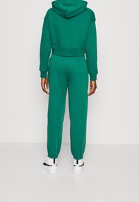 Sweat à capuche vert court avec capuche et manches longues, assorti à un jogging vert. Tissu en coton texturé et poignets côtelés. Baskets blanches.