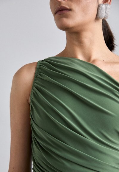 Green draped dress with soft, textured fabric, featuring gathered detailing at the shoulder. Accented by large, silver geometric earrings.