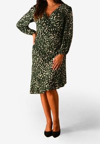 Zeleno-krémové šaty s leopardím vzorem, dlouhými rukávy, zavinovacím designem a rozšířenou sukní, doplněné černými vysokými podpatky.