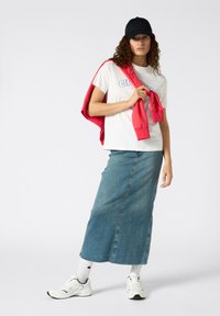 T-shirt blanc avec des lettres bleues, associé à une longue jupe en denim. Sweat-shirt rouge drapé sur les épaules, casquette noire et baskets blanches.