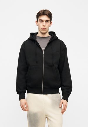 Junger Mann, der eine schwarze Kapuzenjacke mit Reißverschluss, Vorderentaschen und Kordeln trägt, darunter ein graues Hemd und helle beige Hosen, steht vor weißem Hintergrund.