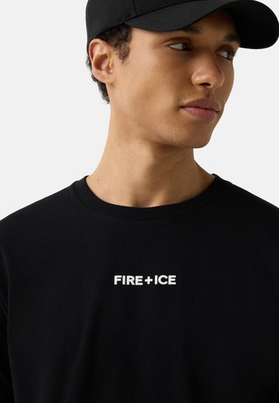 Jeune homme portant une casquette noire et un t-shirt noir avec le texte blanc "FIRE + ICE", regardant vers sa droite sur un fond uni.