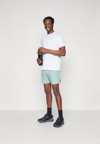 Λευκή αθλητική μπλούζα, μέint πράσινες βερμούδες με νεον πλευρικές λωρίδες, μαύρα παπούτσια, κρα holding black water bottle, standing on a neutral background.