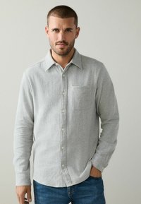 Pilkas flanelinis marškinis su mygtukų užsegimu, viena krūtinės kišenė ir ilgos rankovės. Minkšta tekstūra ir šiek tiek laisvas siluetas.