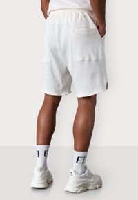 Weiße strukturierte Shorts mit einem elastischen Bund, ausgestattet mit seitlichen Taschen und einer Gesäßtasche. Kombiniert mit weißen Sportschuhen und Socken.