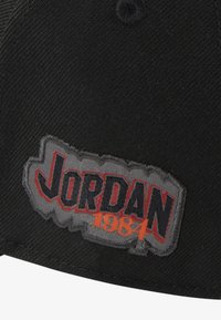 Svart cap med et flerfarget broderi med "JORDAN 1984." Merket har en grå bakgrunn med rød og mørkeblå tekst.