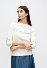 Top bianco a maniche lunghe con scollo a spalle scoperte, trama arricciata sul davanti e maniche aderenti. Abbinato a una gonna di jeans.