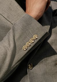 Manche de blazer texturé gris mettant en vedette cinq boutons de couleur claire, avec un gros plan sur le tissage et les détails de couture du tissu.