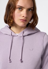 Giovane con capelli corti castani che indossa una felpa leggera di colore viola chiaro con un piccolo logo Champion sul petto, rivolto in avanti contro uno sfondo neutro.