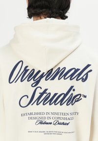 Felpa crema con una grande grafica stampata in blu navy sul retro, che presenta "Originals Studio" e altri dettagli testuali in font eleganti.