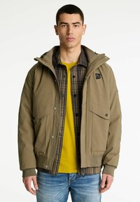 Uomo che indossa una giacca verde oliva sopra una camicia a quadri marrone e una maglietta gialla, abbinata a jeans blu, in piedi su uno sfondo neutro.
