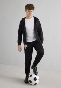 Casaco com fecho éclair preto e calças de fato de treino pretas com o logótipo do AC Milan, combinados com chuteiras de futebol pretas, a estar em cima de uma bola de futebol preta e branca.