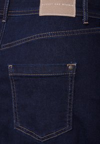 Vaqueros de denim oscuro con un bolsillo trasero que presenta una costura en contraste y una etiqueta cuadrada de tela marrón claro con "STREET ONE STUDIO" grabada.