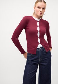 Cardigan lung cu maneci lungi de culoare burgundă, cu dungi, prezentând un guler adânc în V și detalii cu nasturi metalici, asortat cu blugi negri cu picioare largi.