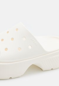 Bijele gumene cipele s ventilacijskim otvorima, teksturiranom potplatom i uzdignutim detaljima. Imaju naziv branda "crocs" utisnut sa strane.