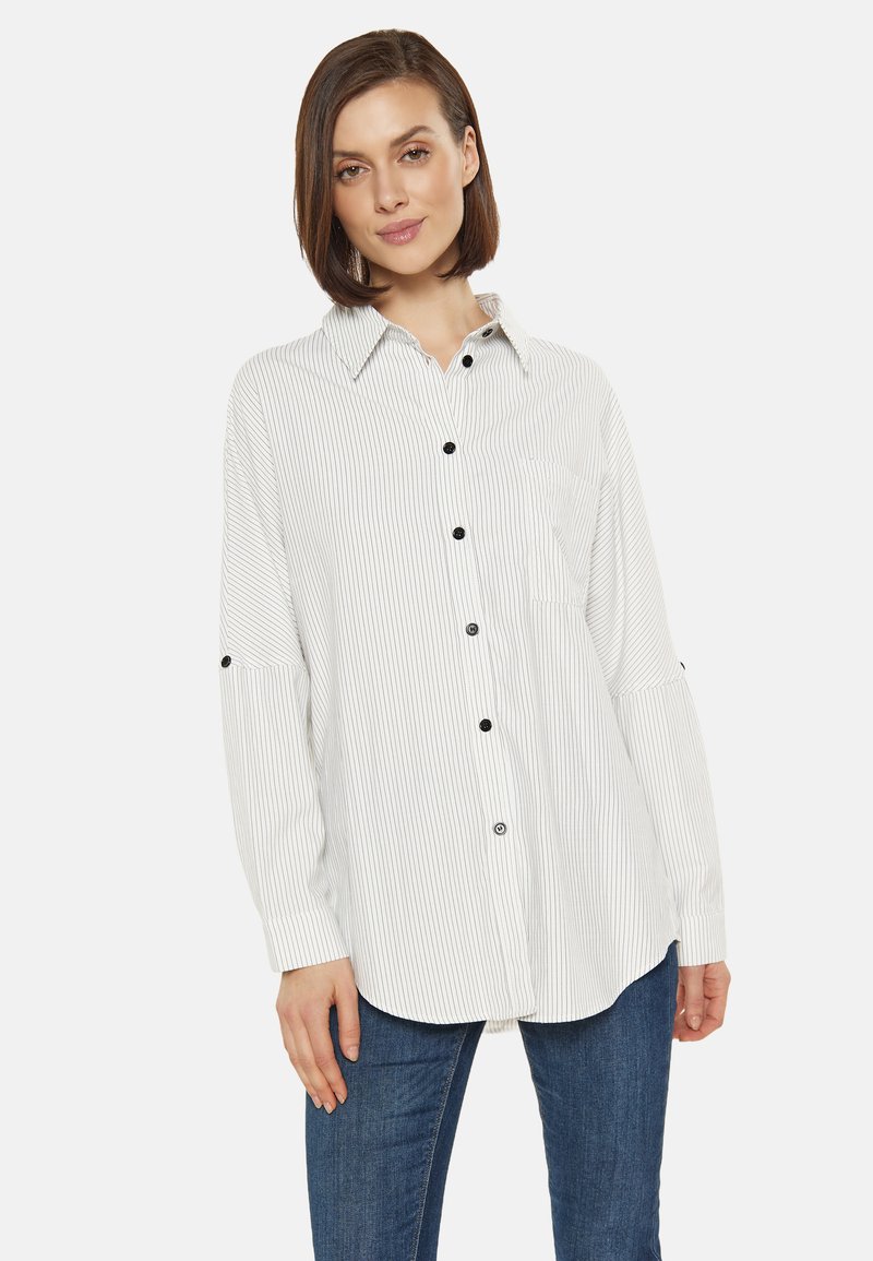 Camisa de botones blanca con rayas y botones negros, con cuello y mangas con tiras enrollables, combinada con jeans azules. Textura suave y corte relajado.