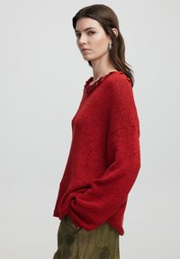 Roter Strickpullover mit lässigem Schnitt, strukturiertem Stoff und geflochtenem Halsausschnitt. Kombiniert mit grünen, gemusterten Hosen.