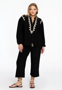Zwarte blouse met verfijnde borduursels langs de halslijn, lange mouwen, gecombineerd met zwarte cropped broek en gestreepte sandalen.
