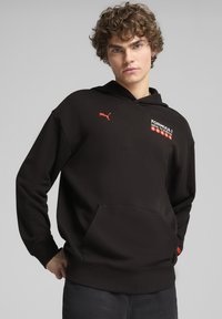 Sudadera negra con bolsillo frontal tipo canguro, presenta el logotipo rojo de Puma y el texto gráfico "Formula 1 Drive to Survive" en el pecho. Tejido suave, corte relajado.