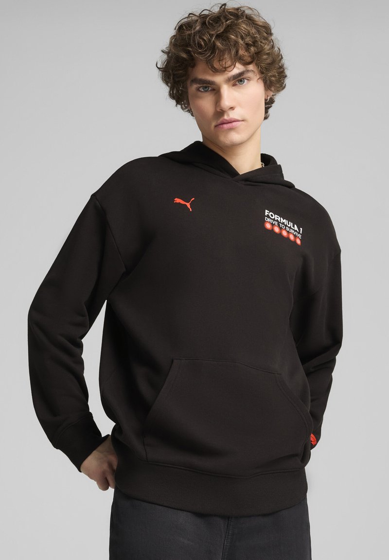 Sudadera negra con bolsillo frontal tipo canguro, presenta el logotipo rojo de Puma y el texto gráfico "Formula 1 Drive to Survive" en el pecho. Tejido suave, corte relajado.