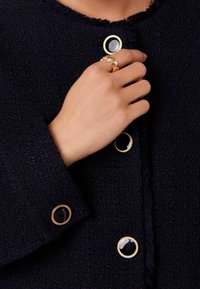 Hand met gouden schelp ring die een donker gestructureerd jack raakt met grote zwart en gouden decoratieve knopen.