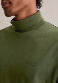 Zeleni rolak od glatke pamučne tkanine, s diskretnim brendiranjem u tamnozelenoj boji sprijeda, s prilagođenim krojem i bez vidljivih šavova.