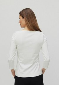 Haut blanc à manches longues avec un décolleté carré, présentant une texture lisse et une coupe ajustée, montré de dos sur un fond uni.