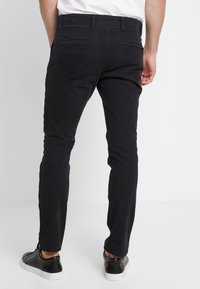 Pantalones slim-fit negros de algodón, con bolsillos traseros y una textura suave. Combinados con zapatillas negras y una camisa blanca.