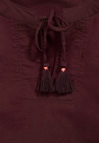 Cordon avec deux pompons sur l'encolure avant d'un vêtement en lin ou coton de couleur bordeaux foncé.