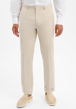 Uomo che indossa pantaloni beige con passanti per cintura, abbinati a scarpe beige senza lacci e una camicia bianca a maniche lunghe.