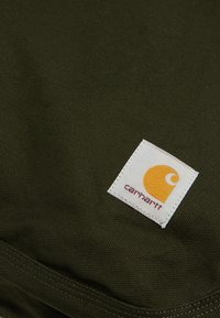 Oliivivihreä kangas, jossa on teksturoitu pinta ja valkoinen suorakulmainen etiketti, jossa on keltainen logo ja "carhartt" maroonin värillä.