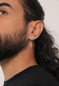 Orecchino a cerchio in argento con un piccolo pendente a croce, caratterizzato da una finitura lucida. Sono visibili capelli ricci e una maglietta scura.